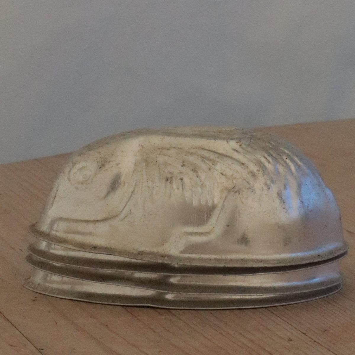 A small silver-toned metal dome-shaped object with engraved detailing, shown resting on a light wood surface against a pale background.