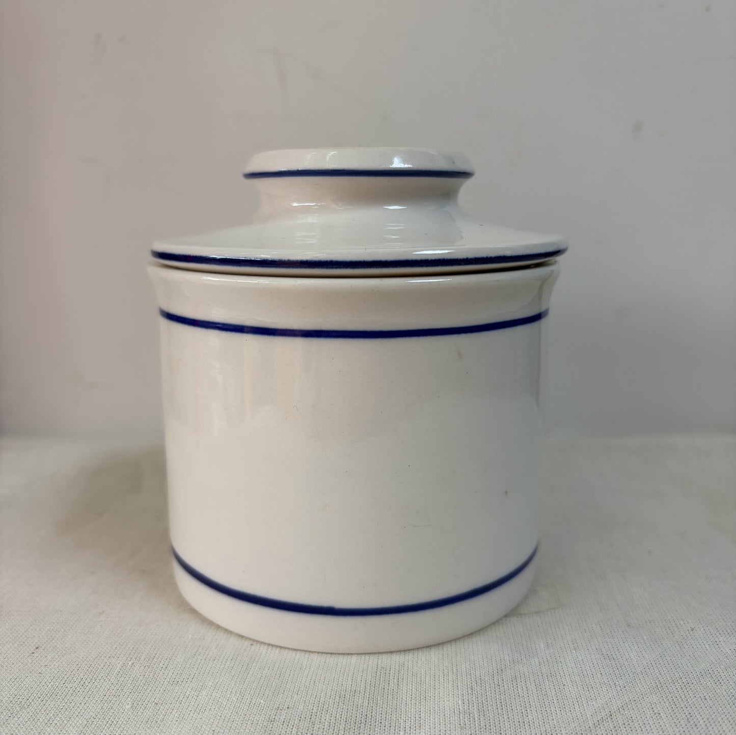 A white ceramic French butterbell or butter crock decorated with two thin blue horizontal stripes.