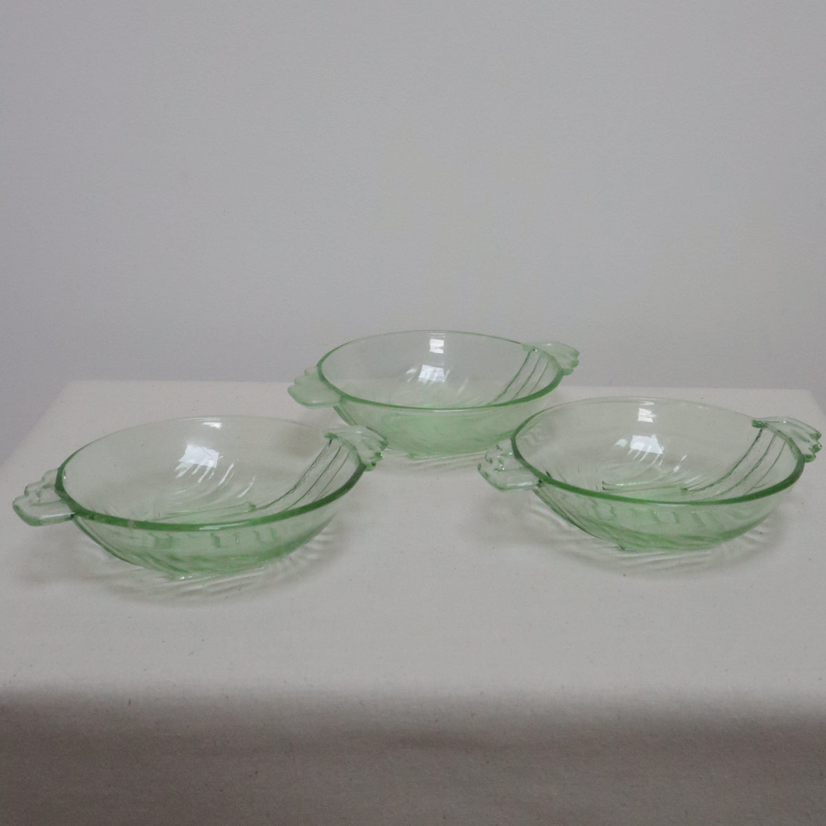 Three small clear glass bowls with light green rims, arranged vertically on a white rectangular surface against a plain light gray wall.