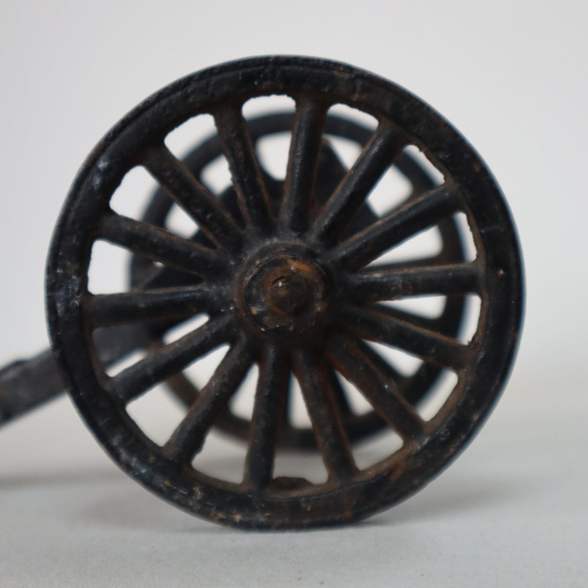 A close-up of a vintage black metal wheel or spoked wheel component with a rustic, weathered finish, set against a plain off-white background.