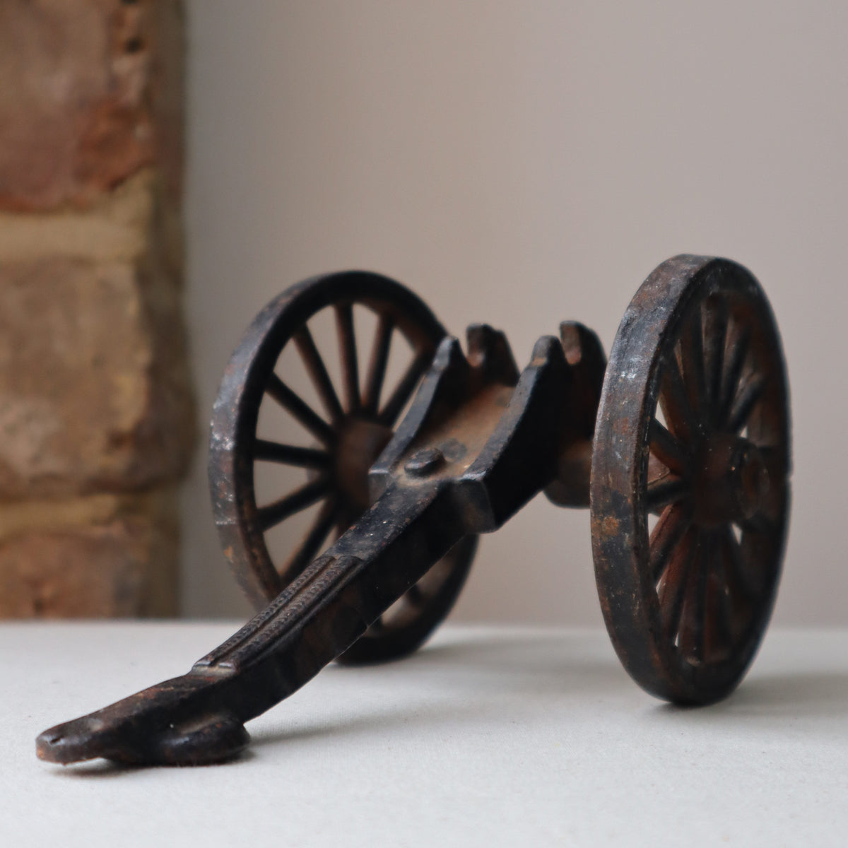 A miniature dark brown wooden toy cannon with two spoked wheels, displayed on a light surface with a blurred brick column and neutral background.