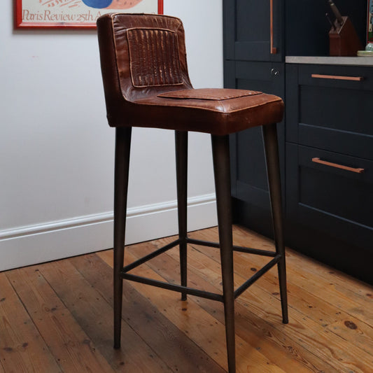A brown leather bar stool with a dark metal frame and legs, positioned on a wooden floor next to a dark kitchen cabinet.