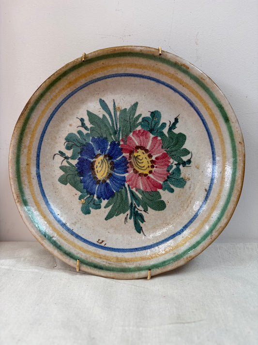 A vintage ceramic plate with a hand-painted floral design featuring blue and pink flowers with green leaves, surrounded by concentric painted stripes.