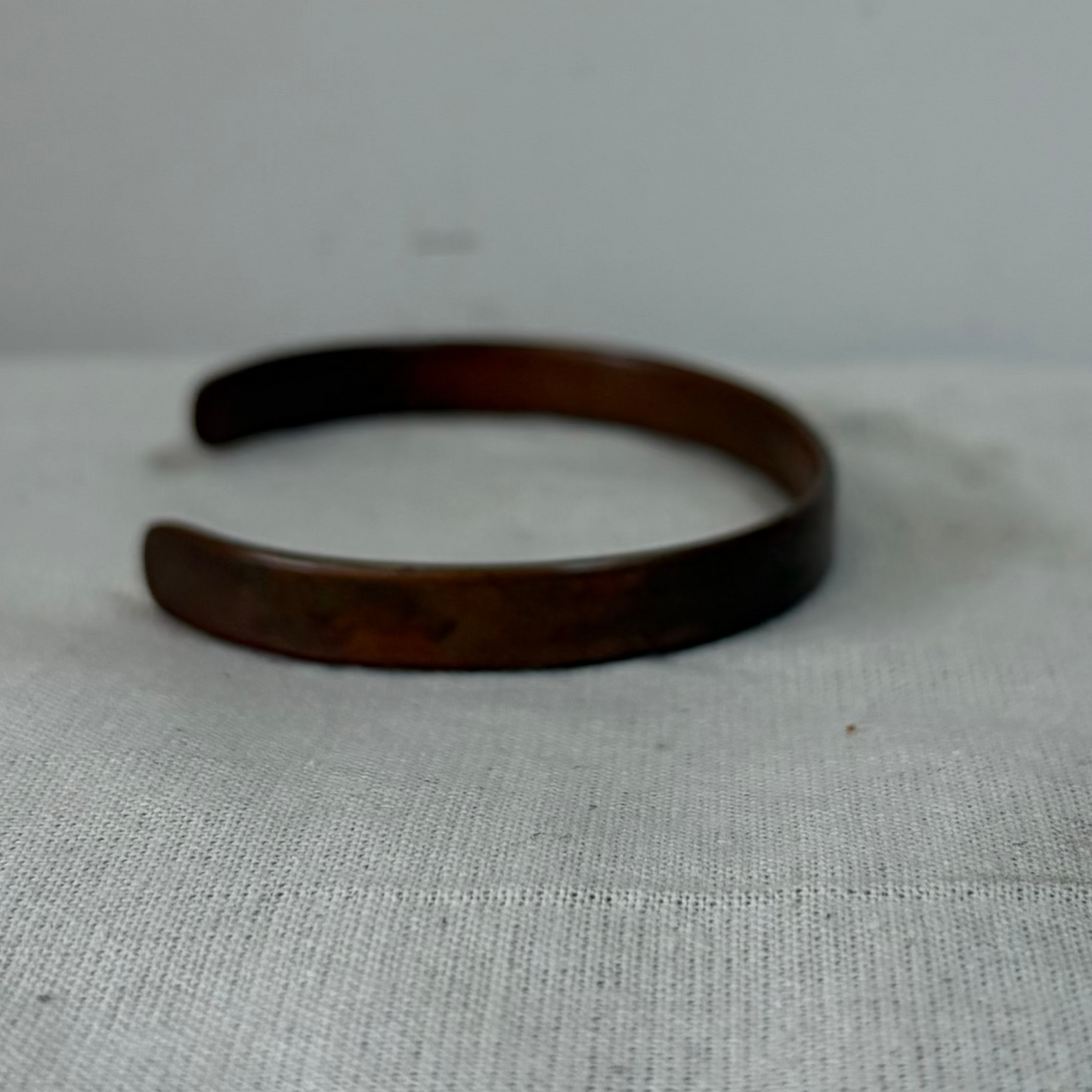 A dark brown open cuff bracelet, likely made of wood or a wood-like material, displayed on a light gray fabric surface against a matching light gray background.