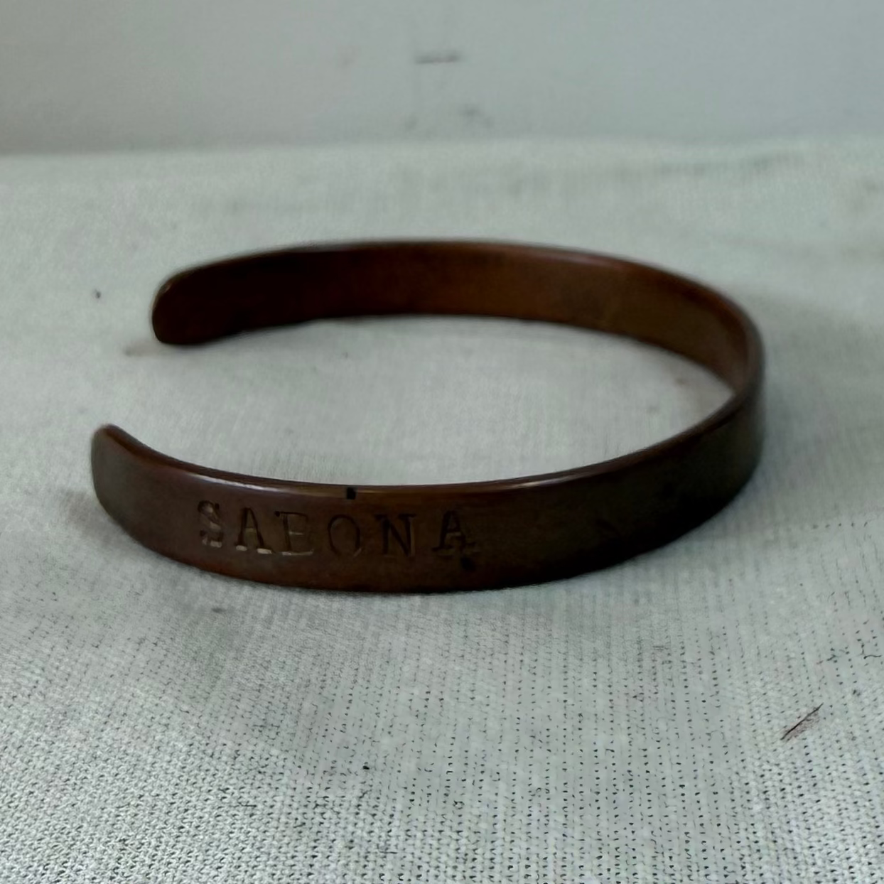 A dark brown wooden cuff bracelet with a natural split opening, resting on a textured off-white fabric surface against a plain light gray background.