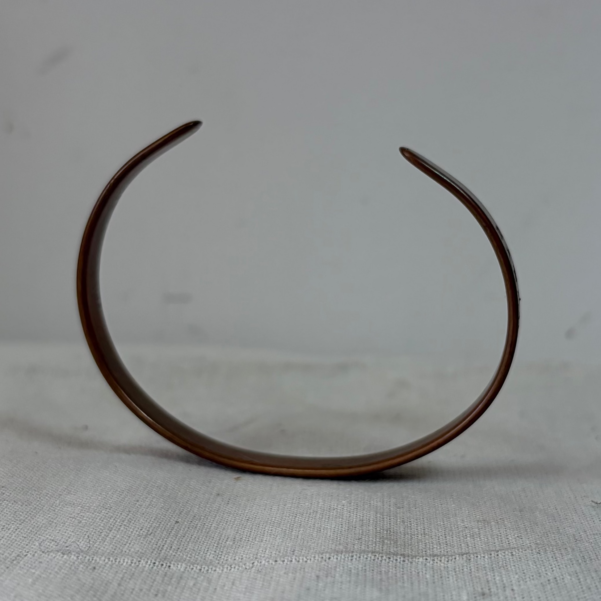 A minimalist, open-ended curved metal cuff bracelet in a dark bronze finish, standing upright on a light gray textured fabric surface against a neutral gray background.