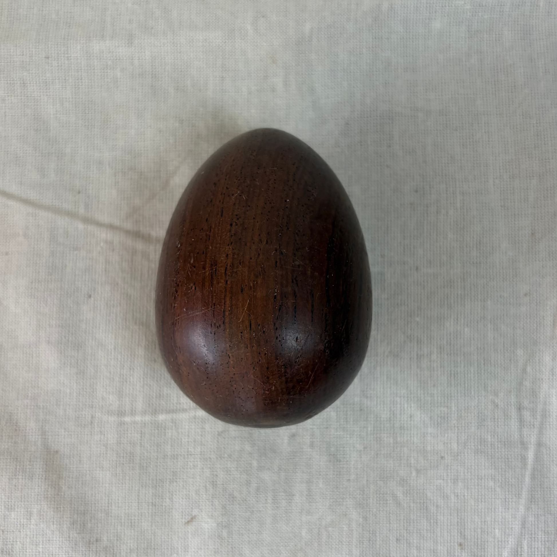 A small, smooth, wooden egg with a glossy finish, displayed against a draped light beige linen fabric background.