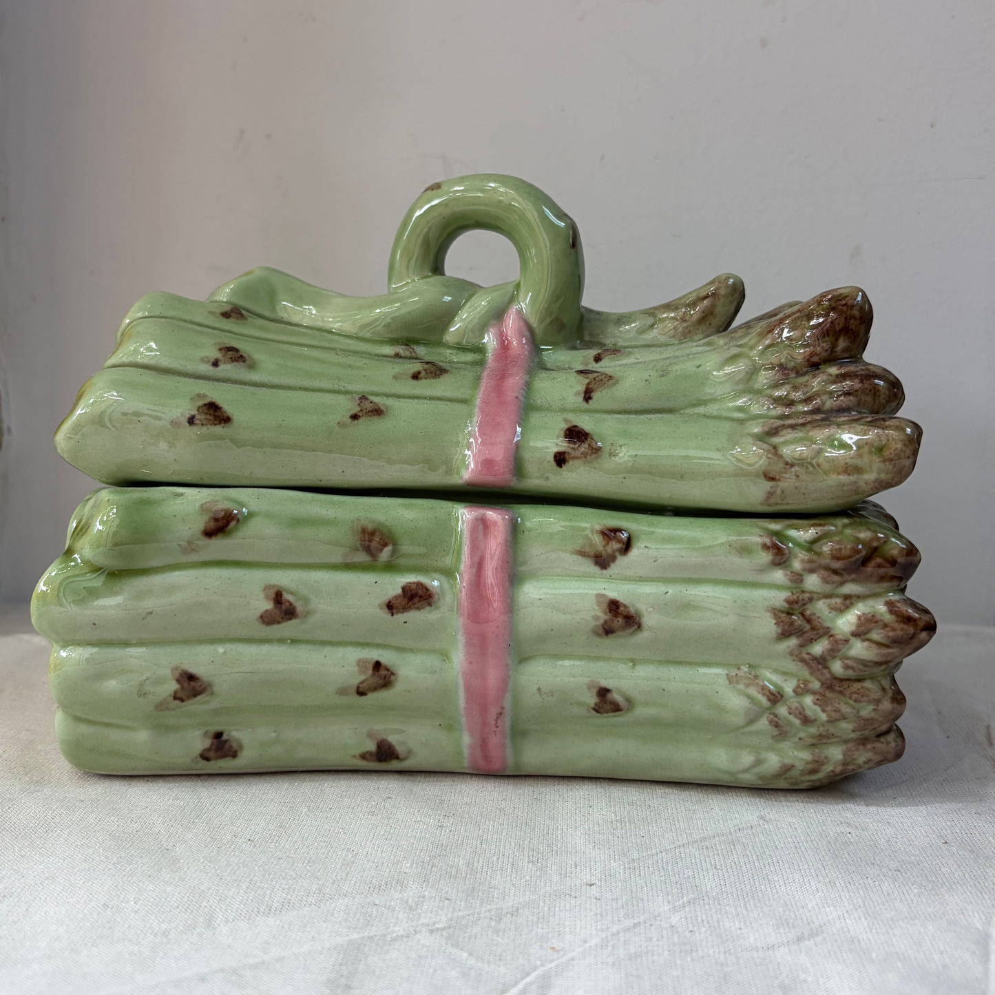 A ceramic asparagus-shaped container with a matching lid, featuring a glossy light green glaze, brown speckles, and a pink band around the center, displayed on a white fabric surface.