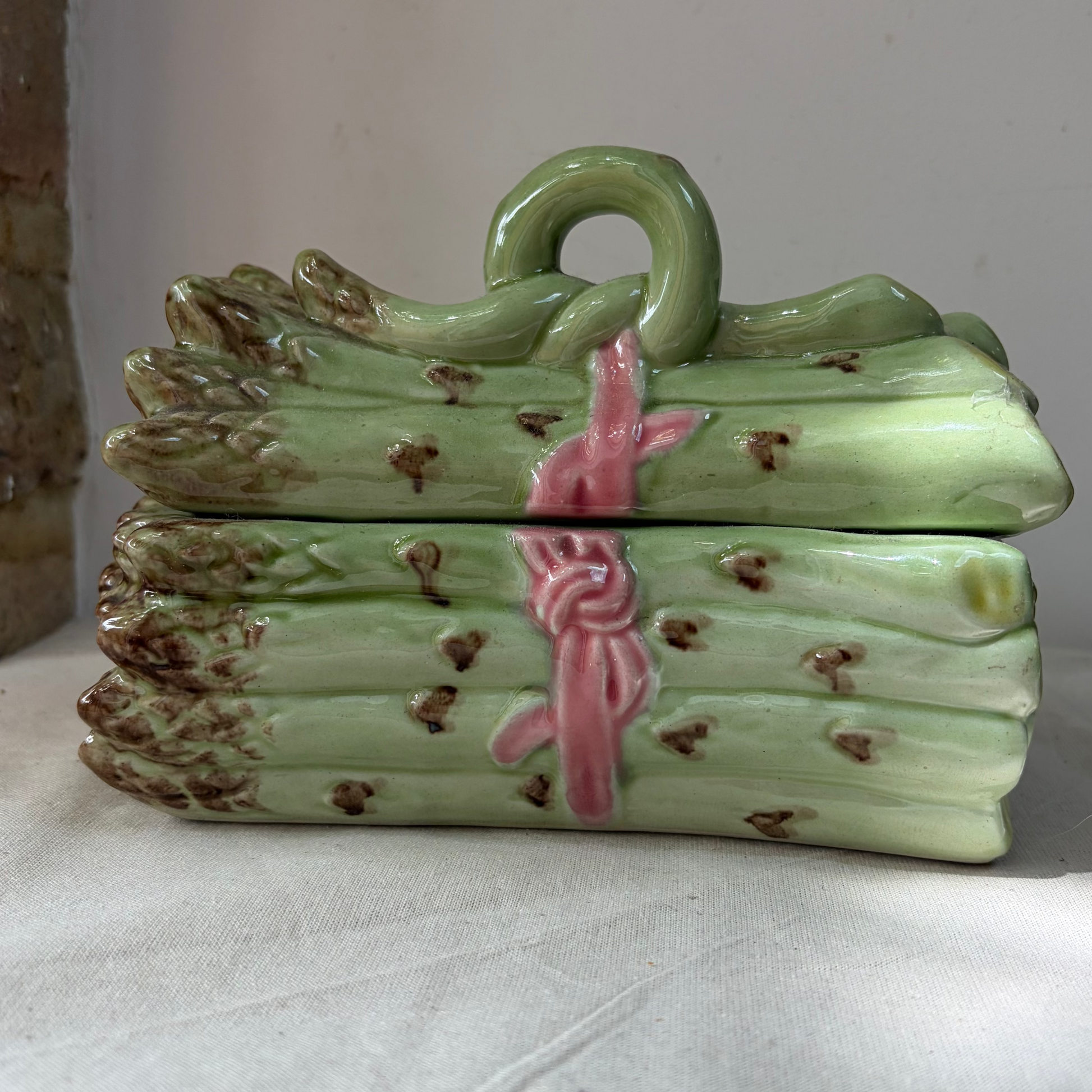 A decorative ceramic box shaped like a bundle of asparagus spears, with a light green glazed body, brown speckled tips, and a pink ribbon tied around the centre, sitting on a white fabric surface.