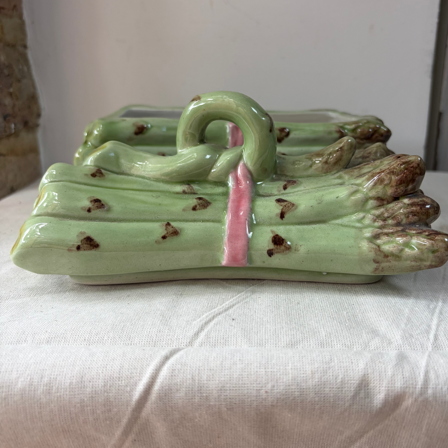 A ceramic asparagus-shaped decorative object with a glossy light green glaze, brown speckled tips, and a pink center band, displayed on a white fabric-covered surface.