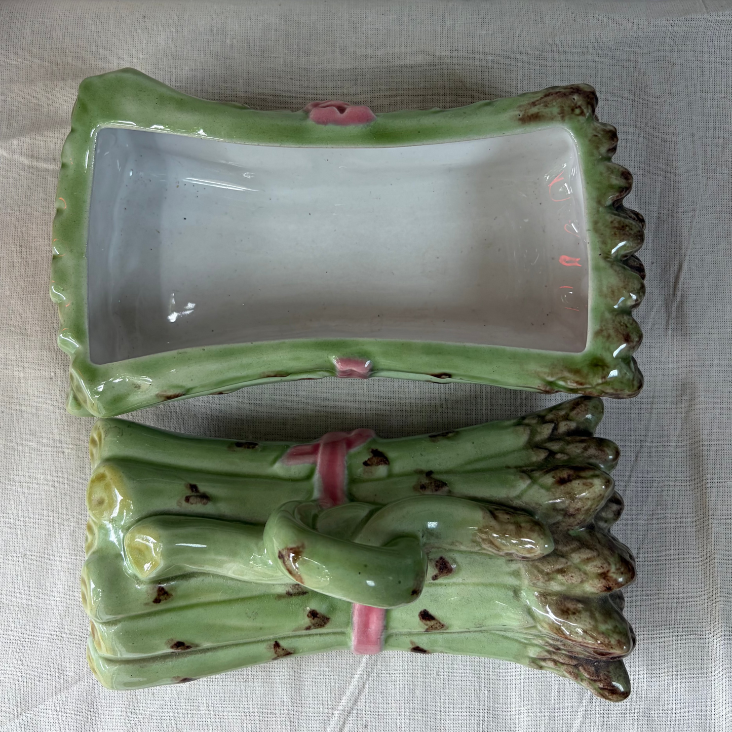 A ceramic asparagus dish, featuring a rectangular green glazed tray with a raised asparagus stem detail tied with a pink ribbon, displayed on a light fabric surface.